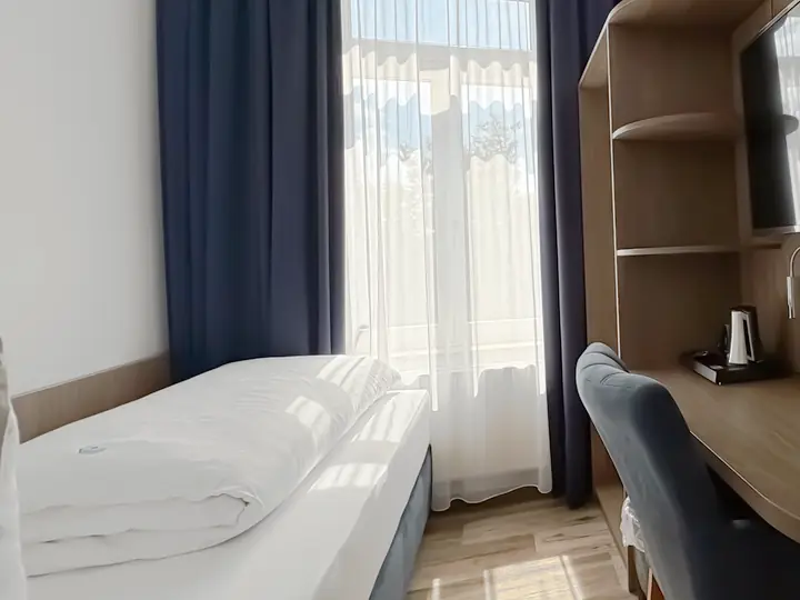 Einzelzimmer mit Bett am Fenster, Schreibtisch und blauem Sessel