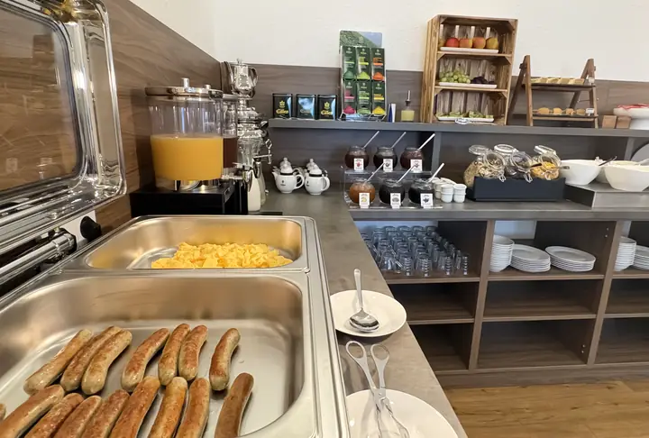 Warmhaltebuffet mit Würstchen und Rührei, daneben Saftspender, Gläser und Teller