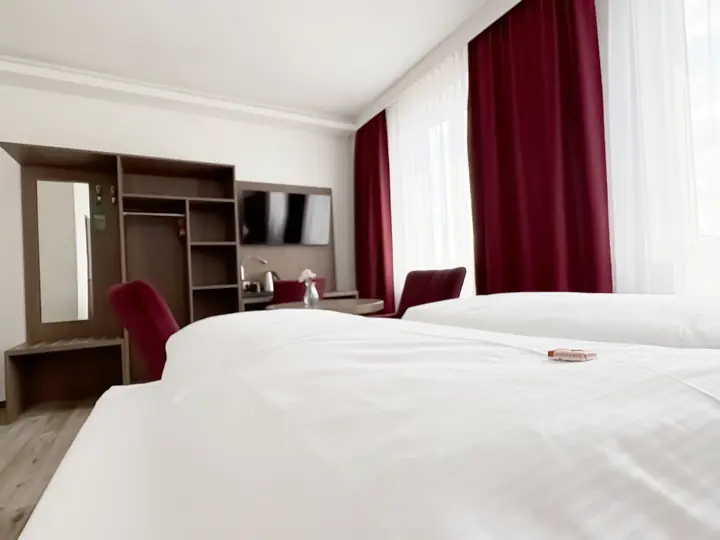 Doppelzimmer in Weinrot: Betten, offener Schrank mit Spiegel, Fernseher und zwei weinrote Sessel